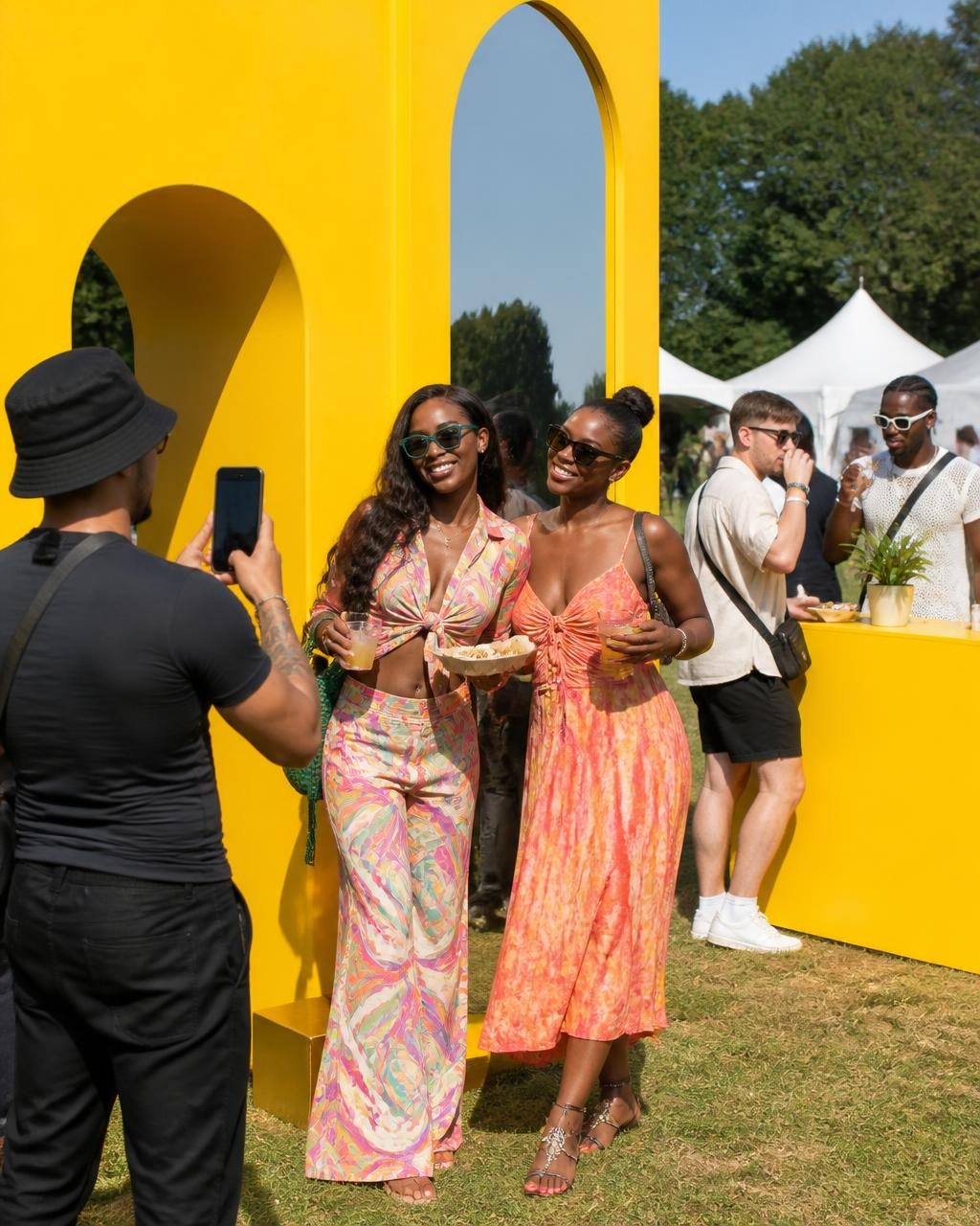 Two friends posing for a photo in front of a yellow arched festival installation while guests sample drinks nearby